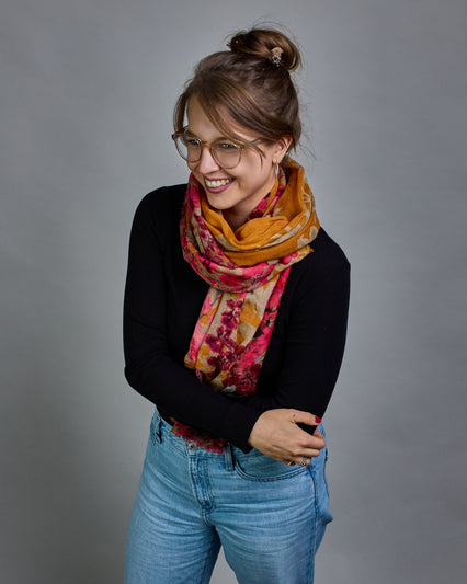 Ein Schal aus 75 % Wolle und 25 % Modal mit den Maßen 70 × 180 cm. Je nach Bild ist er locker um den Nacken gelegt oder einmal gewickelt, um eine lockere Schlaufe um den Hals zu haben, oder zweimal gewickelt, um das Maximum an Wärme zu bekommen. An den Kanten ist er leicht gefranst. Die Hintergrundfarbe des Schals ist Okker. Von den schmalen Kanten des Schals kommen die Blumen in Richtung Mitte. Die Blüten sind in Rosa-, Lila- und Weißtönen gestaltet. #2
