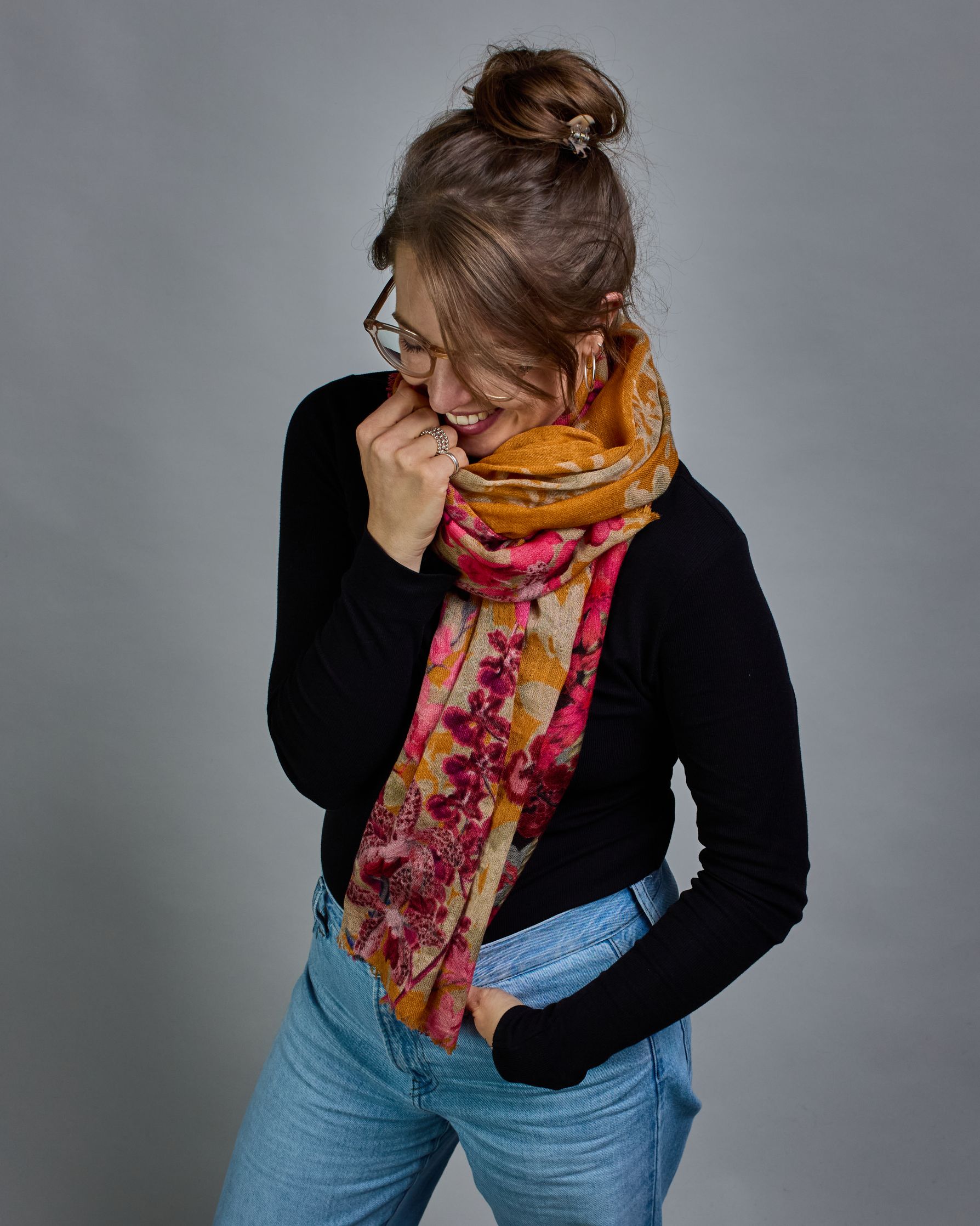 Ein Schal aus 75 % Wolle und 25 % Modal mit den Maßen 70 × 180 cm. Je nach Bild ist er locker um den Nacken gelegt oder einmal gewickelt, um eine lockere Schlaufe um den Hals zu haben, oder zweimal gewickelt, um das Maximum an Wärme zu bekommen. An den Kanten ist er leicht gefranst. Die Hintergrundfarbe des Schals ist Okker. Von den schmalen Kanten des Schals kommen die Blumen in Richtung Mitte. Die Blüten sind in Rosa-, Lila- und Weißtönen gestaltet. #3