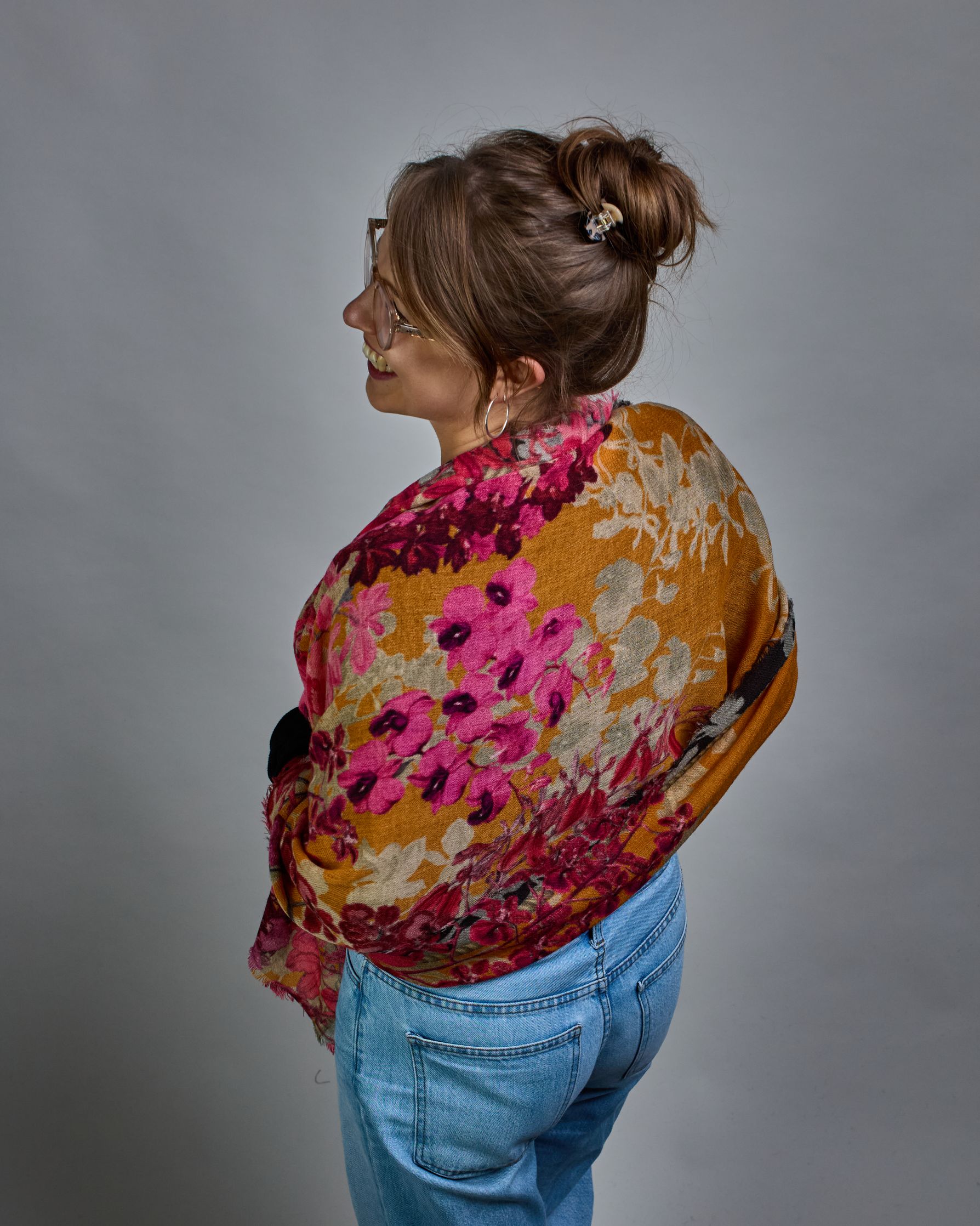 Ein Schal aus 75 % Wolle und 25 % Modal mit den Maßen 70 × 180 cm. Je nach Bild ist er locker um den Nacken gelegt oder einmal gewickelt, um eine lockere Schlaufe um den Hals zu haben, oder zweimal gewickelt, um das Maximum an Wärme zu bekommen. An den Kanten ist er leicht gefranst. Die Hintergrundfarbe des Schals ist Okker. Von den schmalen Kanten des Schals kommen die Blumen in Richtung Mitte. Die Blüten sind in Rosa-, Lila- und Weißtönen gestaltet. #4