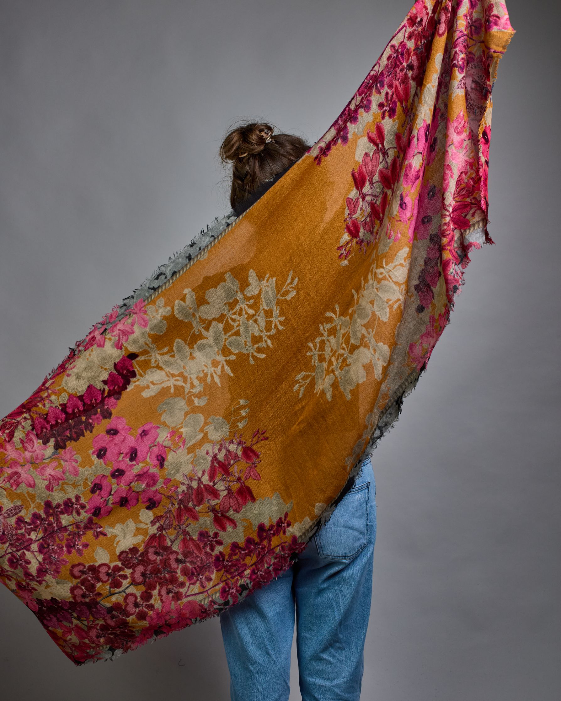 Ein Schal aus 75 % Wolle und 25 % Modal mit den Maßen 70 × 180 cm. Je nach Bild ist er locker um den Nacken gelegt oder einmal gewickelt, um eine lockere Schlaufe um den Hals zu haben, oder zweimal gewickelt, um das Maximum an Wärme zu bekommen. An den Kanten ist er leicht gefranst. Die Hintergrundfarbe des Schals ist Okker. Von den schmalen Kanten des Schals kommen die Blumen in Richtung Mitte. Die Blüten sind in Rosa-, Lila- und Weißtönen gestaltet. #5