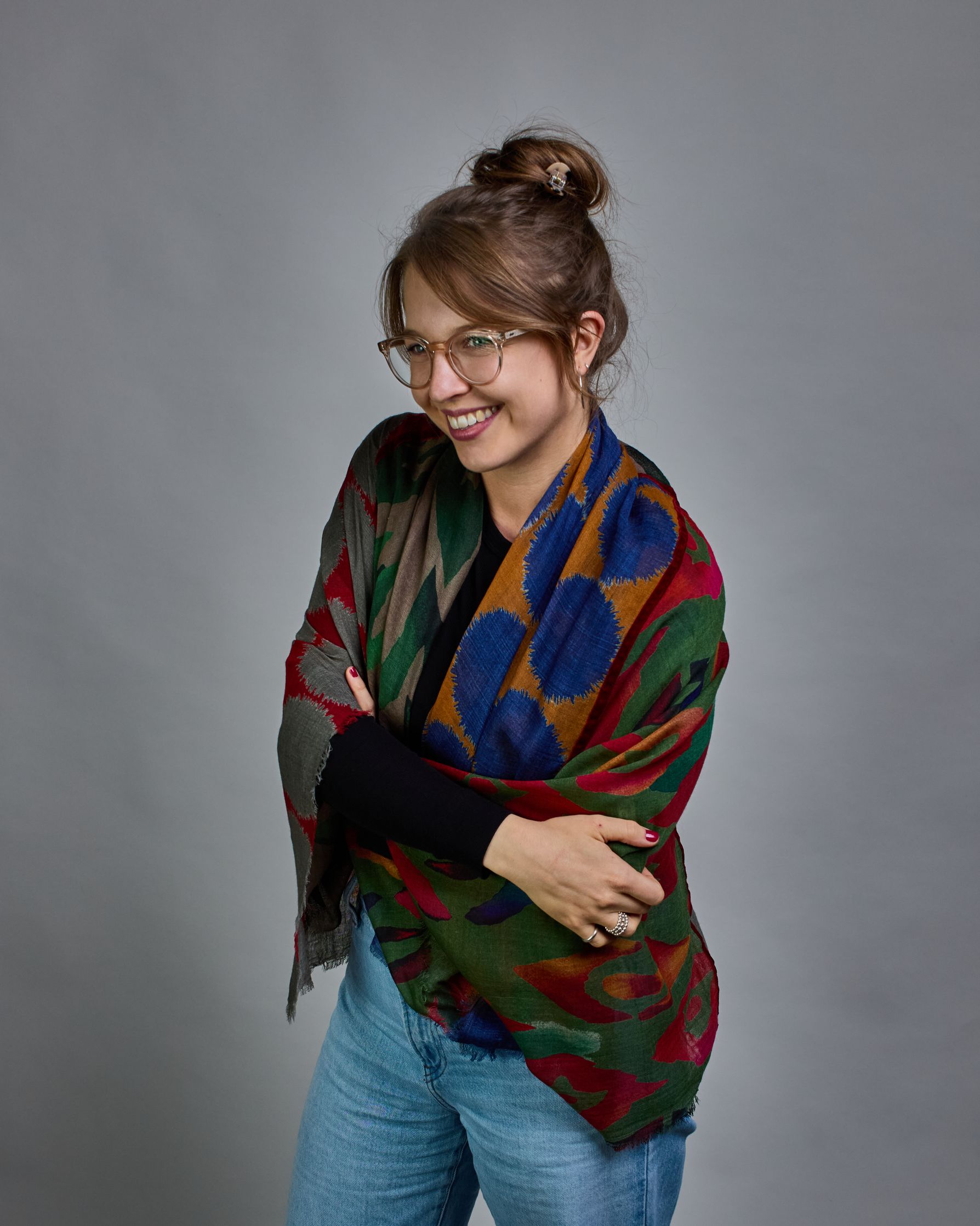 Ein Schal aus 90 % Wolle und 10% Seide. Je nach Bild ist er locker um den Nacken gelegt oder einmal gewickelt, um eine lockere Schlaufe um den Hals zu haben, oder zweimal gewickelt, um das Maximum an Wärme zu bekommen. An den Kanten ist er leicht gefranst. Das Design ist in 4 gleichgroße Quadrate aufgeteilt die jeweils ein anderes Muster hat. Die Quadrate sind mit einem aufgenähtem Rotem Samtband unterteilt. Eines der Quadrate hat als Grundfarbe Okker und darauf befinden sich fransige Kreise in Blau. Im nä…