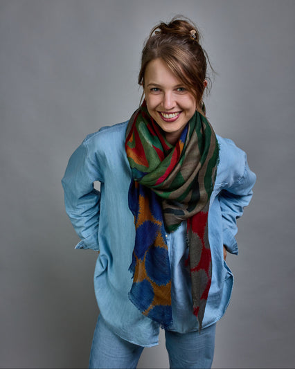 Ein Schal aus 90 % Wolle und 10% Seide. Je nach Bild ist er locker um den Nacken gelegt oder einmal gewickelt, um eine lockere Schlaufe um den Hals zu haben, oder zweimal gewickelt, um das Maximum an Wärme zu bekommen. An den Kanten ist er leicht gefranst. Das Design ist in 4 gleichgroße Quadrate aufgeteilt die jeweils ein anderes Muster hat. Die Quadrate sind mit einem aufgenähtem Rotem Samtband unterteilt. Eines der Quadrate hat als Grundfarbe Okker und darauf befinden sich fransige Kreise in Blau. Im nä…