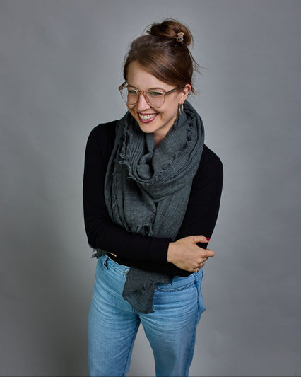 Ein anthrazitfarbener Schal aus 100 % Wolle mit den Maßen 100x180 cm. Je nach Bild ist er locker um den Hals gelegt oder einmal gewickelt, um eine Schlaufe um den Hals zu haben. Der Schal ist einfärbig und hat an den Kanten leichte Fransen. Die organische Struktur verleiht dem Schal ein lässiges, zeitloses Design. #1