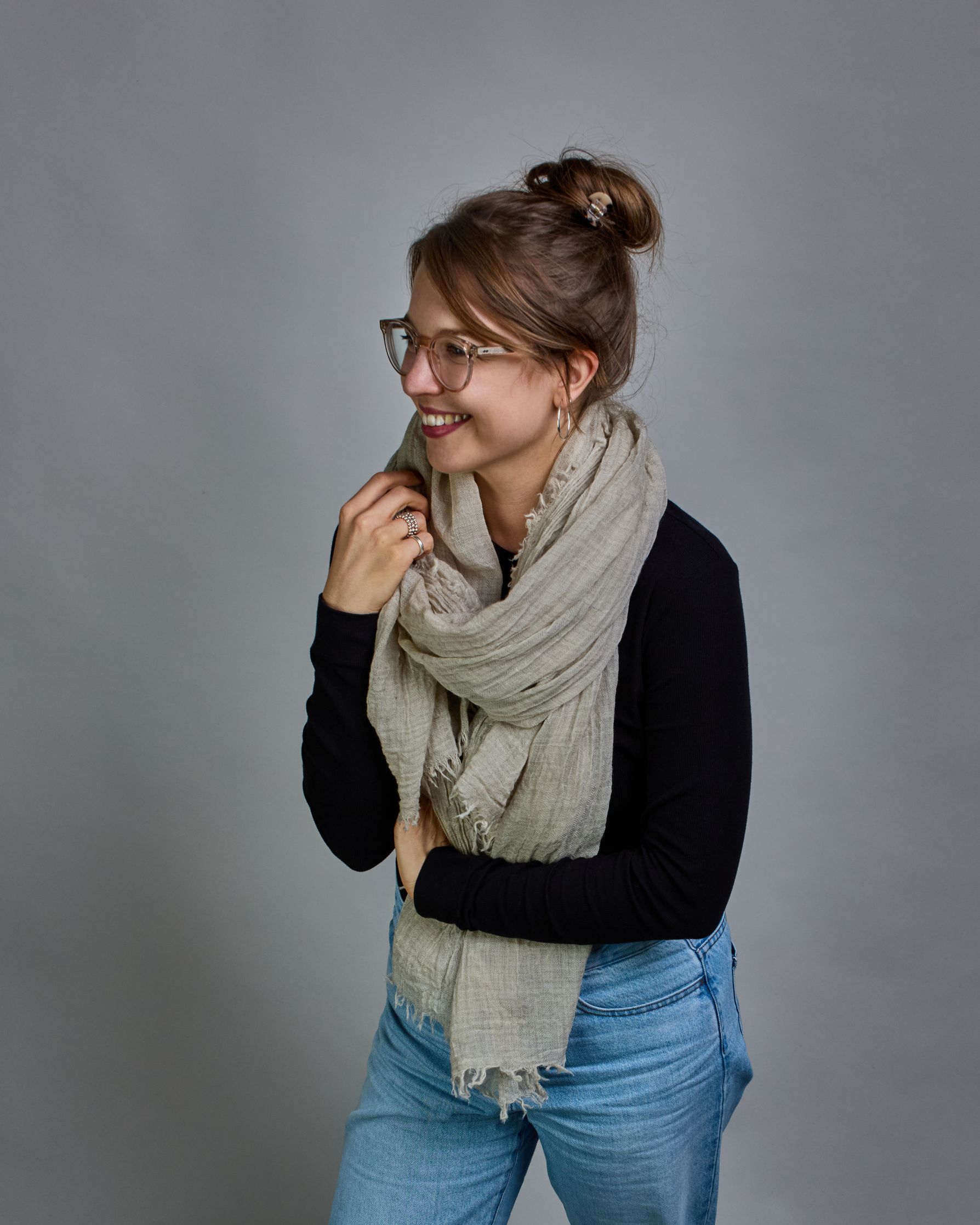 Ein beiger Schal aus 100 % Wolle mit den Maßen 100x180 cm. Je nach Bild ist er locker um den Hals gelegt oder einmal gewickelt, um eine Schlaufe um den Hals zu haben. Der Schal ist einfärbig und hat an den Kanten leichte Fransen. Die organische Struktur verleiht dem Schal ein lässiges, zeitloses Design. #3