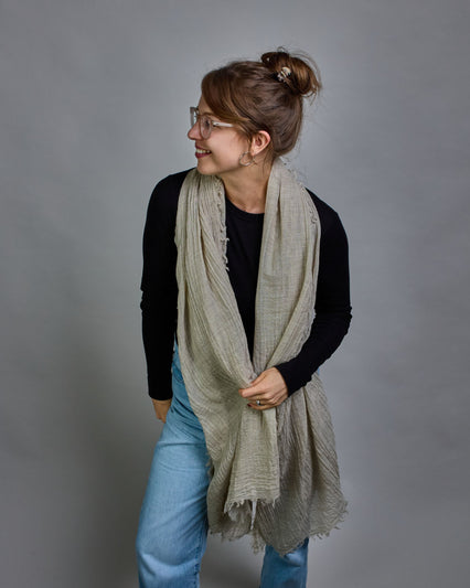 Ein beiger Schal aus 100 % Wolle mit den Maßen 100x180 cm. Je nach Bild ist er locker um den Hals gelegt oder einmal gewickelt, um eine Schlaufe um den Hals zu haben. Der Schal ist einfärbig und hat an den Kanten leichte Fransen. Die organische Struktur verleiht dem Schal ein lässiges, zeitloses Design. #5