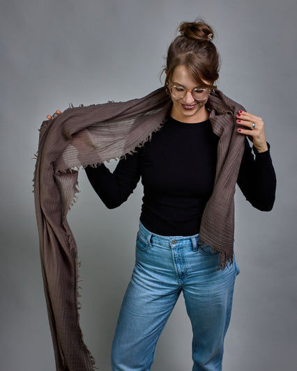 Ein Schal aus 100 % Wolle in einem kaltem Braunton mit den Maßen 100x180 cm. Je nach Bild ist er locker um den Hals gelegt oder einmal gewickelt, um eine Schlaufe um den Hals zu haben. Der Schal ist einfärbig und hat an den Kanten leichte Fransen. Die organische Struktur verleiht dem Schal ein lässiges, zeitloses Design. #4