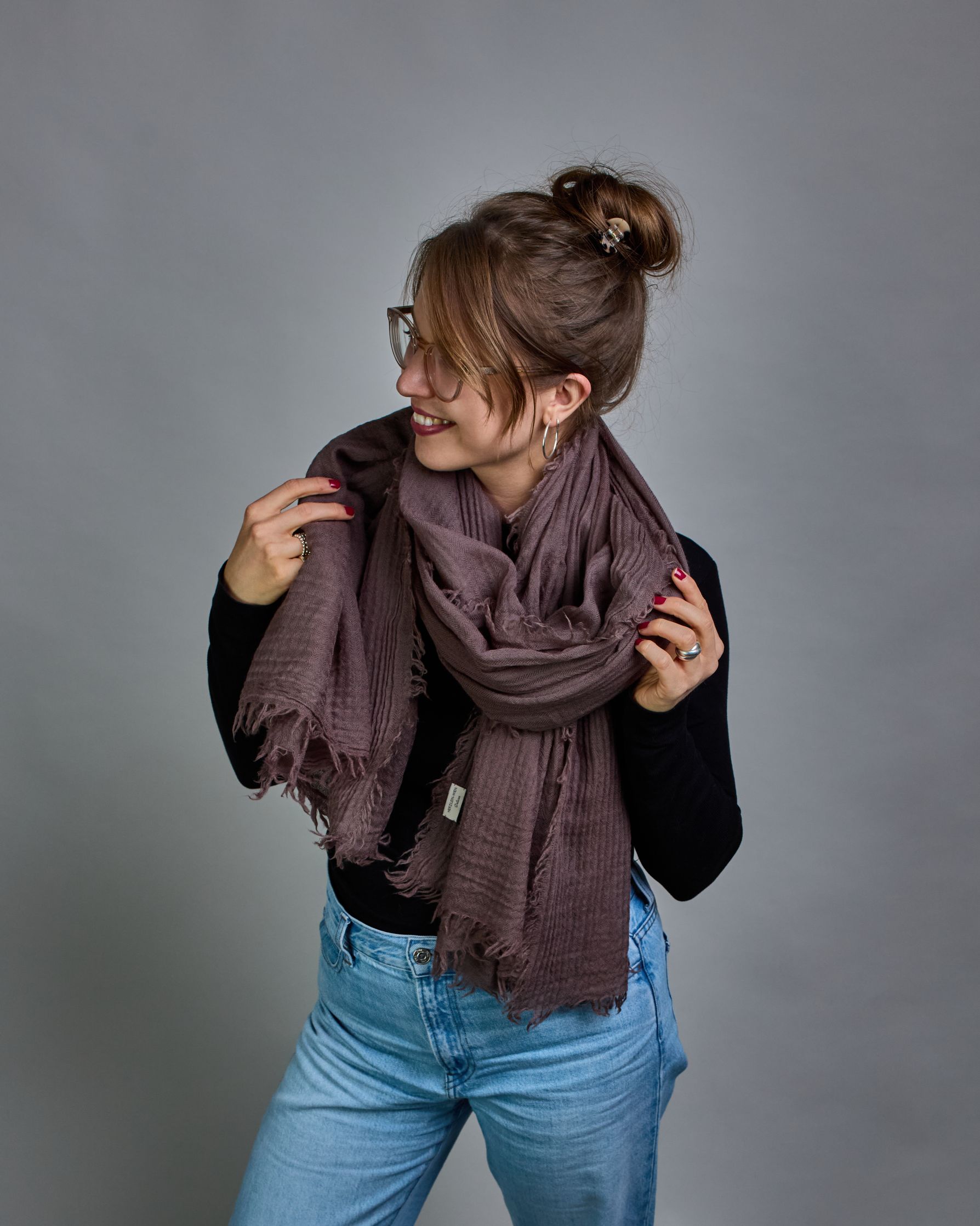 Ein Schal aus 100 % Wolle in einem warmen Braunton mit den Maßen 100x180 cm. Je nach Bild ist er locker um den Hals gelegt oder einmal gewickelt, um eine Schlaufe um den Hals zu haben. Der Schal ist einfärbig und hat an den Kanten leichte Fransen. Die organische Struktur verleiht dem Schal ein lässiges, zeitloses Design. #3