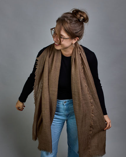 Ein cognacfarbener Schal aus 100 % Wolle mit den Maßen 100x180 cm. Je nach Bild ist er locker um den Hals gelegt oder einmal gewickelt, um eine Schlaufe um den Hals zu haben. Der Schal ist einfärbig und hat an den Kanten leichte Fransen. Die organische Struktur verleiht dem Schal ein lässiges, zeitloses Design. #4