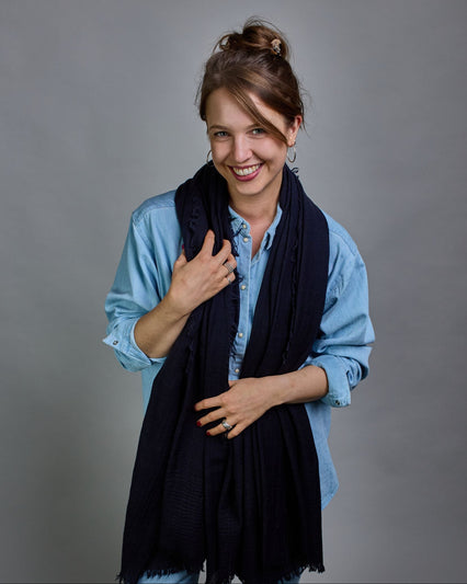 Ein dunkelblauer Schal aus 100 % Wolle mit den Maßen 100x180 cm. Je nach Bild ist er locker um den Hals gelegt oder einmal gewickelt, um eine Schlaufe um den Hals zu haben. Der Schal ist einfärbig und hat an den Kanten leichte Fransen. Die organische Struktur verleiht dem Schal ein lässiges, zeitloses Design. #6