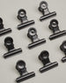 Ein zehnerset von Metall-Clips in mattem schwarz mit einem Loch in den Greifarmen an denen man sie oben öffnen kann. #16