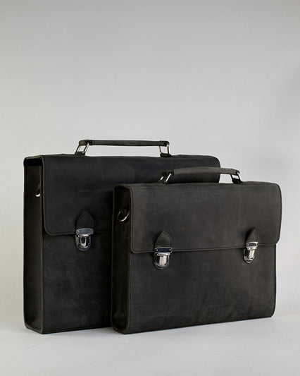 Schwarze Aktentasche aus Hunterleder mit Tragegriff, abnehmbarem Schulterriemen, Metallverschlüssen sowie Innenfächer. Das Lederist anfangs matt und entwickelt mit der Zeit eine individuelle Patina. Logo Prägung unter der Lasche mit zwei Metallverschlüssen in klassischem Schultaschen-Stil. #17