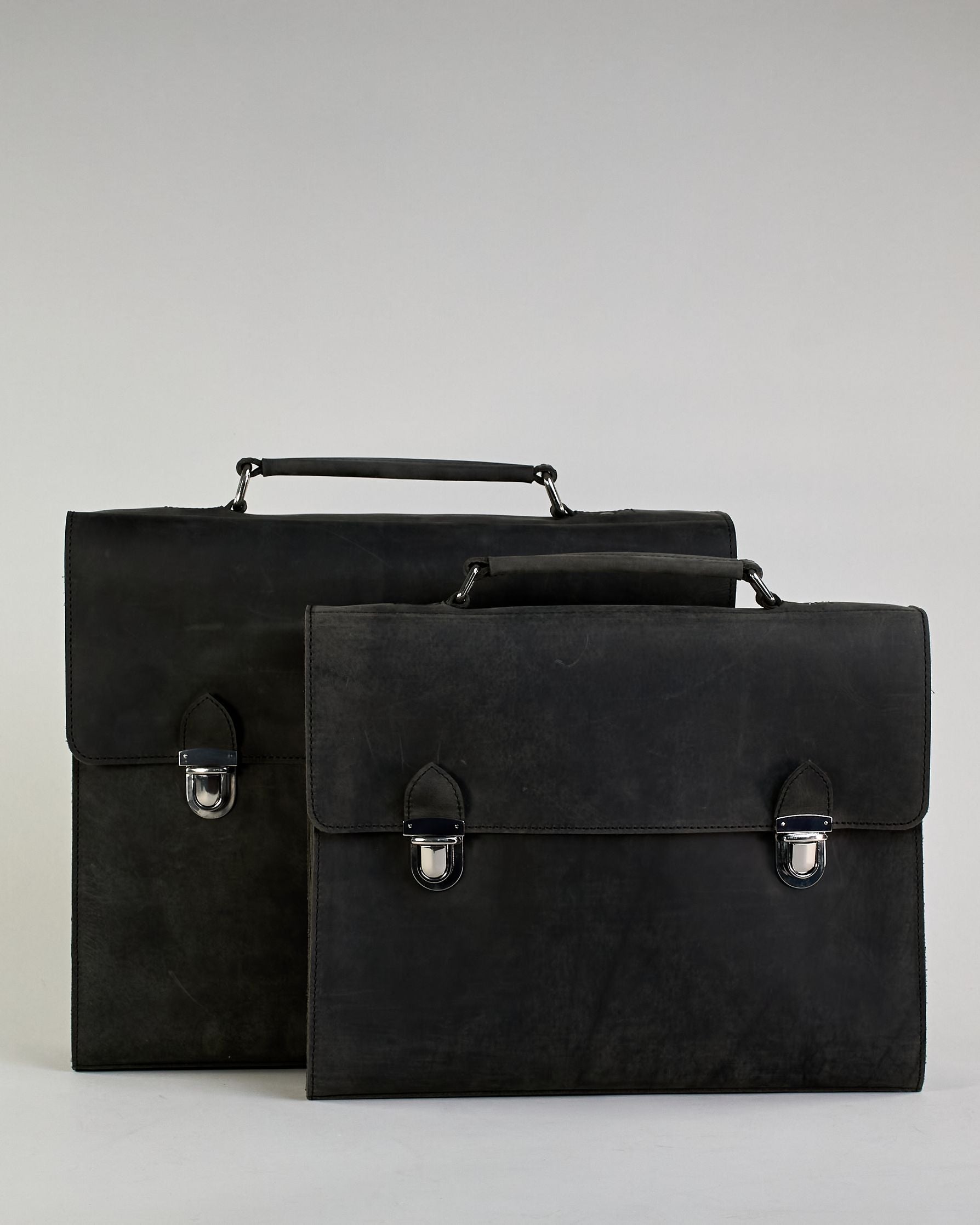 Schwarze Aktentasche aus Hunterleder mit Tragegriff, abnehmbarem Schulterriemen, Metallverschlüssen sowie Innenfächer. Das Lederist anfangs matt und entwickelt mit der Zeit eine individuelle Patina. Logo Prägung unter der Lasche mit zwei Metallverschlüssen in klassischem Schultaschen-Stil. #18