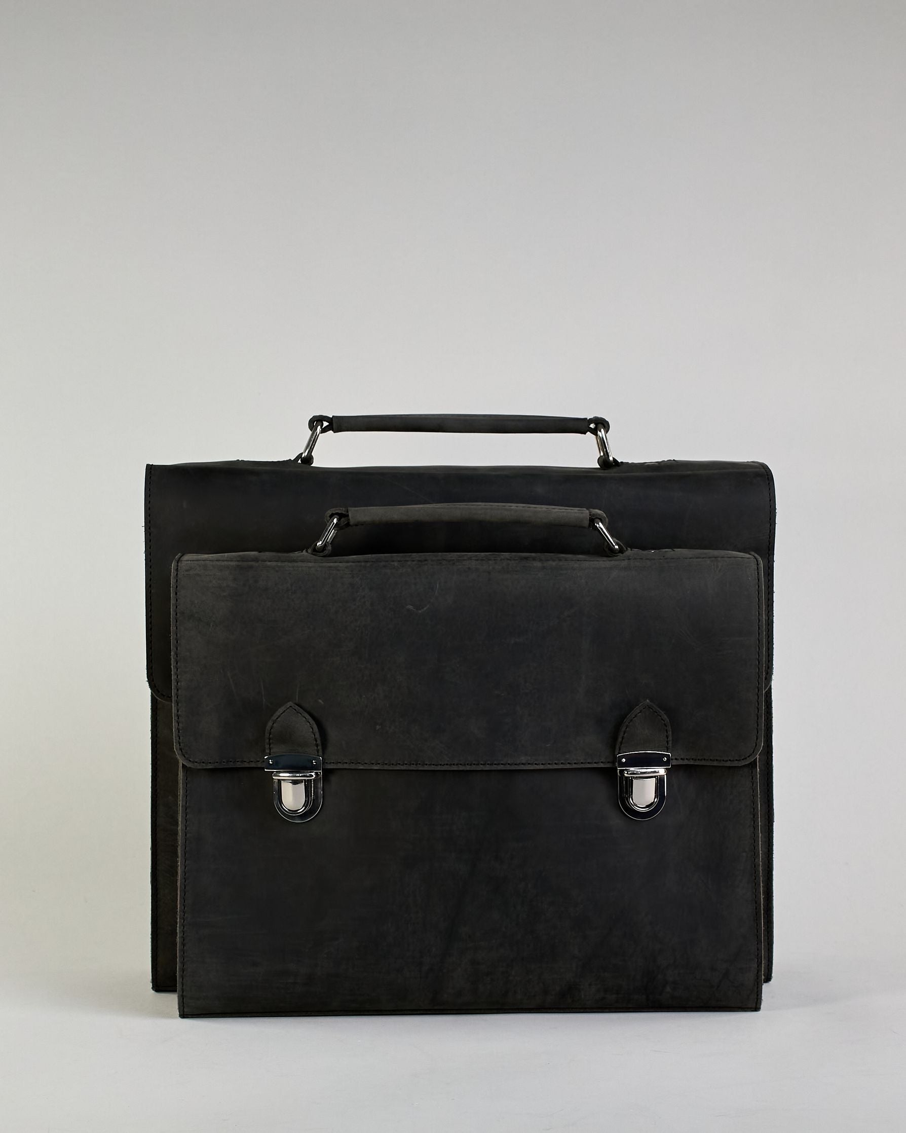 Schwarze Aktentasche aus Hunterleder mit Tragegriff, abnehmbarem Schulterriemen, Metallverschlüssen sowie Innenfächer. Das Lederist anfangs matt und entwickelt mit der Zeit eine individuelle Patina. Logo Prägung unter der Lasche mit zwei Metallverschlüssen in klassischem Schultaschen-Stil. #20