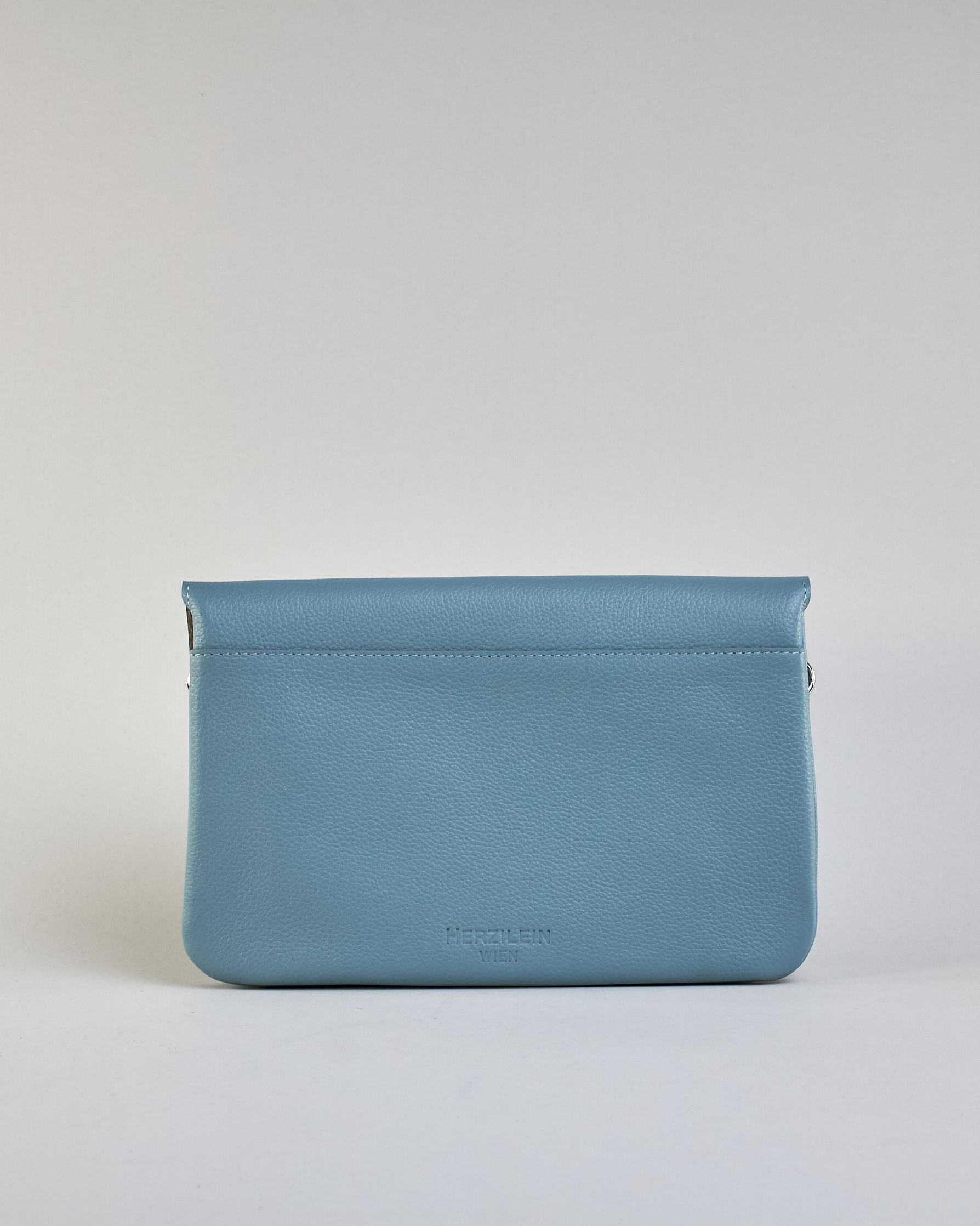 Einfärbige kleine Crossbody-Tasche aus genarbtem Rindleder mit Umschlaglasche und silbernem Druckknopf. Die Tasche enthält innen ein Zippfach, ein offenes Fach sowie ein schmales Zwischenfach. Auf der Rückseite ist eine Logoprägung "Herzilein-Wien" #43