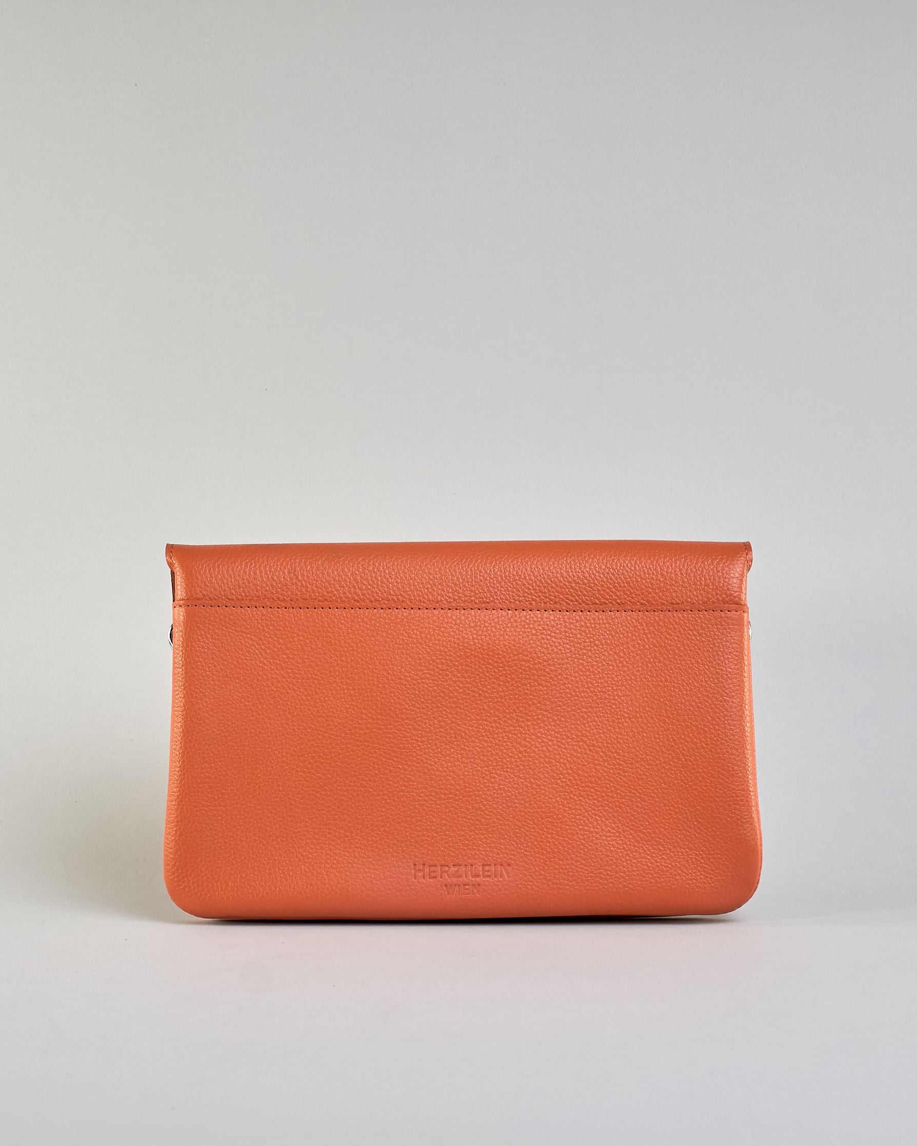 Einfärbige kleine Crossbody-Tasche aus genarbtem Rindleder mit Umschlaglasche und silbernem Druckknopf. Die Tasche enthält innen ein Zippfach, ein offenes Fach sowie ein schmales Zwischenfach. Auf der Rückseite ist eine Logoprägung "Herzilein-Wien" #49