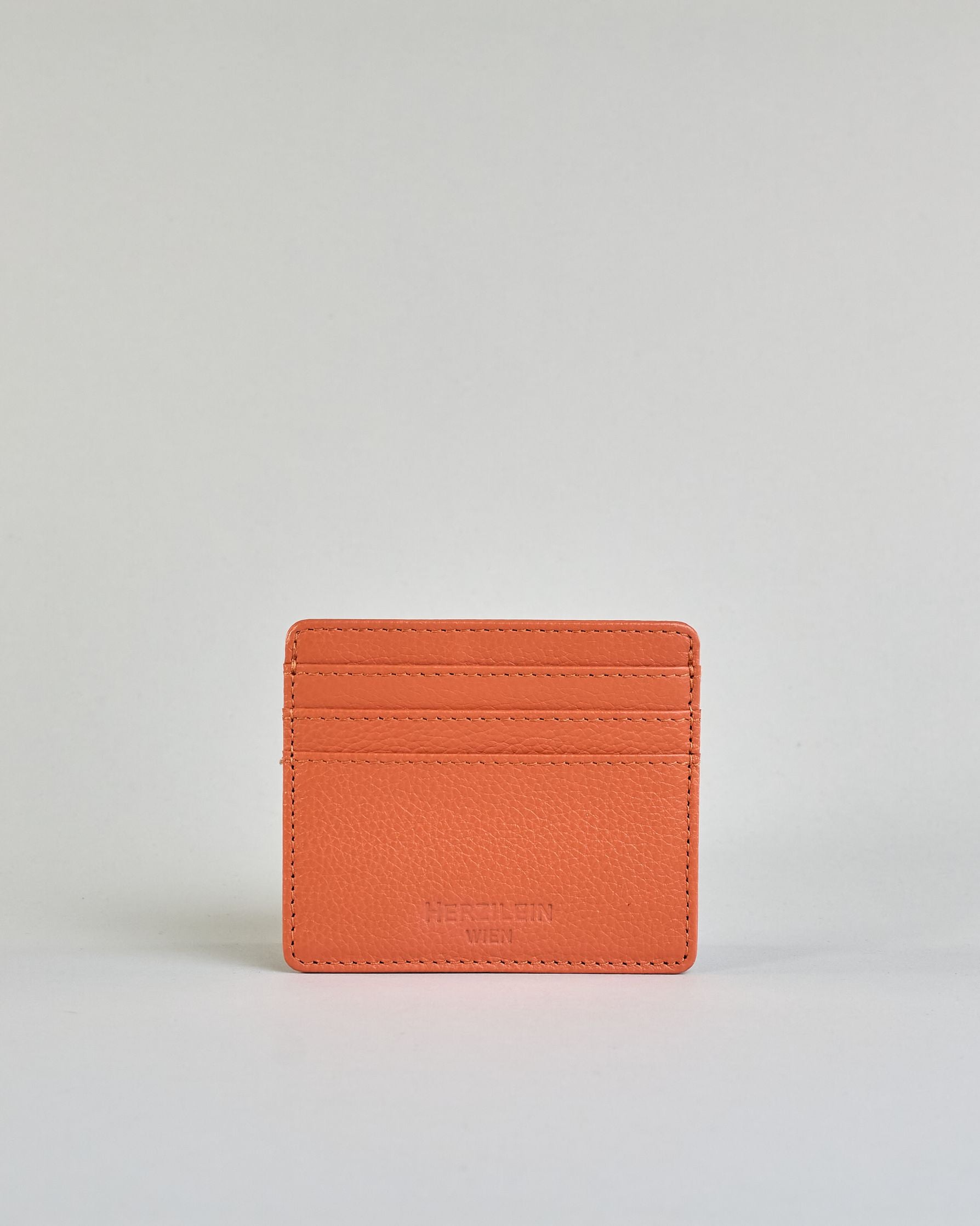 Geldbörse aus genarbtem Rindleder mit Umschlaglasche und silbernem Druckknopf. Die Geldbörse enthält innen ein Zippfach, ein offenes Fach sowie ein schmales Zwischenfach. Auf der Rückseite ist eine Logoprägung "Herzilein-Wien"